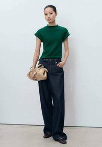 Green short-sleeve top, dark wide-leg denim pants, and a cream handbag; model stands against a neutral background with flat shoes.