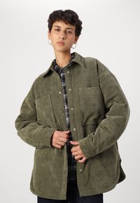 Camisa jaqueta de cotel verde-azeitona com dois bolsos frontais, botões de pressão e um corte descontraído, sobre uma camisa xadrez preta e cinza.