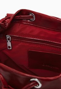 Roter, glanzender Tascheninneraum mit einem Reißverschlussfach, metallischen Beschlägen und einer glatten Innenauskleidung. Enthält ein Lederetikett mit eingraviertem Text.