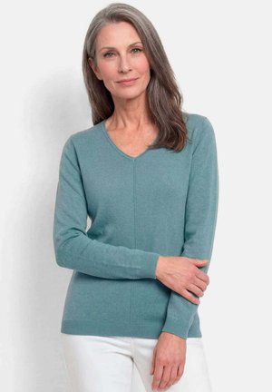 Femme aux longs cheveux gris portant un pull à col en V de couleur sarcelle et un pantalon blanc, debout avec un bras croisé sur son corps devant un fond uni.