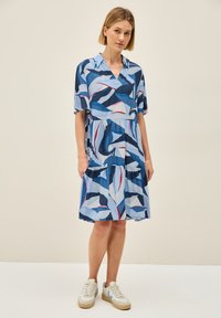 Kurzärmliges, kragenloses Kleid mit einem gestuften, gerüschten Saum. Es zeigt ein blaues Blättermuster auf einem hellblauen Hintergrund. Getragen mit weißen Sneakers.