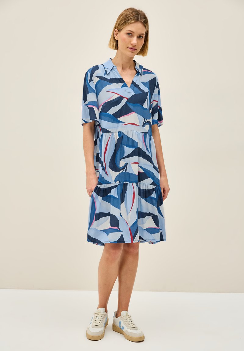 Kurzärmliges, kragenloses Kleid mit einem gestuften, gerüschten Saum. Es zeigt ein blaues Blättermuster auf einem hellblauen Hintergrund. Getragen mit weißen Sneakers.