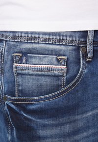 Jeans in denim di un blu scuro con una finitura testurizzata, caratterizzati da una piccola tasca posteriore con cuciture rosse e bianche e un'etichetta con logo.