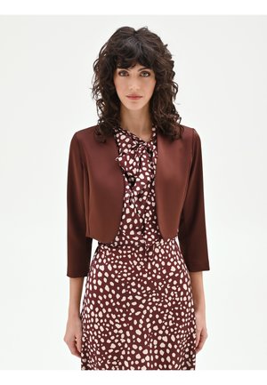 Donna con capelli ricci e scuri che indossa una giacca corta marrone sopra un vestito marrone e bianco a macchie con scollo a fiocco, in piedi davanti a uno sfondo semplice.