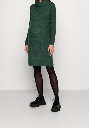 Gravid person iført en grøn strikket sweaterkjole, sorte strømpebukser og sorte chunky snørestøvler, stående foran en ensfarvet baggrund.