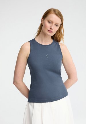 Femme aux cheveux roux et lisses portant un haut sans manches bleu foncé moulant et une jupe blanche, debout devant un fond blanc uni.