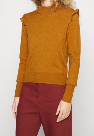 Femme portant un pull à manches longues jaune moutarde avec des détails froncés aux épaules et un pantalon taille haute marron contre un fond uni.