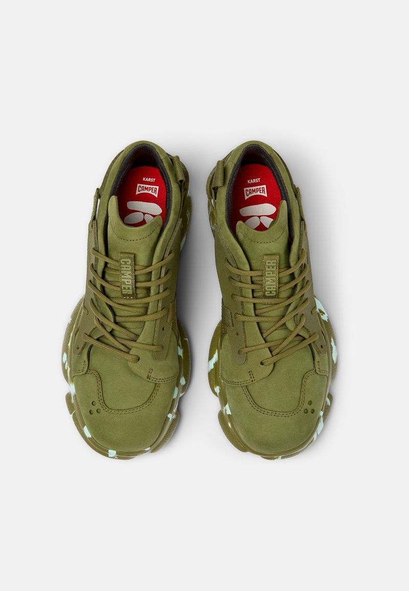 Olivgrüne Sportschuhe mit Obermaterial aus Wildleder, Schnürverschluss, gepolstertem Schaft und reflektierenden Akzenten auf der klobigen Gummisohle. Rote Einlegesohlen sichtbar.