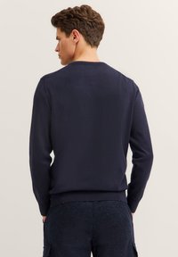 Mann mit kurzem braunen Haar, der einen schlichten dunkelblauen Pullover und dunkel gemusterte Hosen trägt, mit dem Rücken zu einem beigen Hintergrund.
