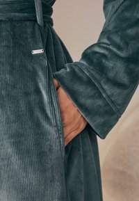 Tmavozelený zamatový župan s vertikálnou rebrovanou textúrou, vybavený opaskom v páse a bočnými vreckami. Dizajn dopĺňa strieborná značka značky.