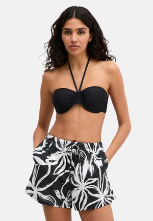 Vrouw met lang donker haar draagt een zwarte strapless bikini top met halterband en zwart-witte korte broek met bloemenpatroon, met handen in de zakken.