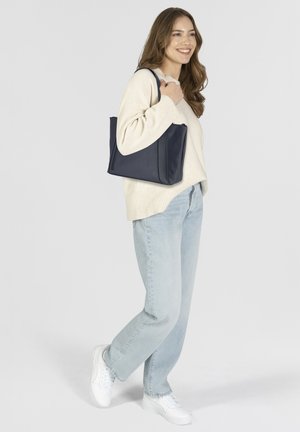 Sac en cuir navy avec une texture lisse et un haut arrondi, présentant un design minimaliste et des poignées doubles, porté au-dessus d'un pull crème.