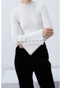 Biały prążkowany top z długim rękawem, kołnierzem ozdobionym perłami oraz rozkloszowanymi rękawami z wycięciami i perłowymi zdobieniami na krawędziach.
