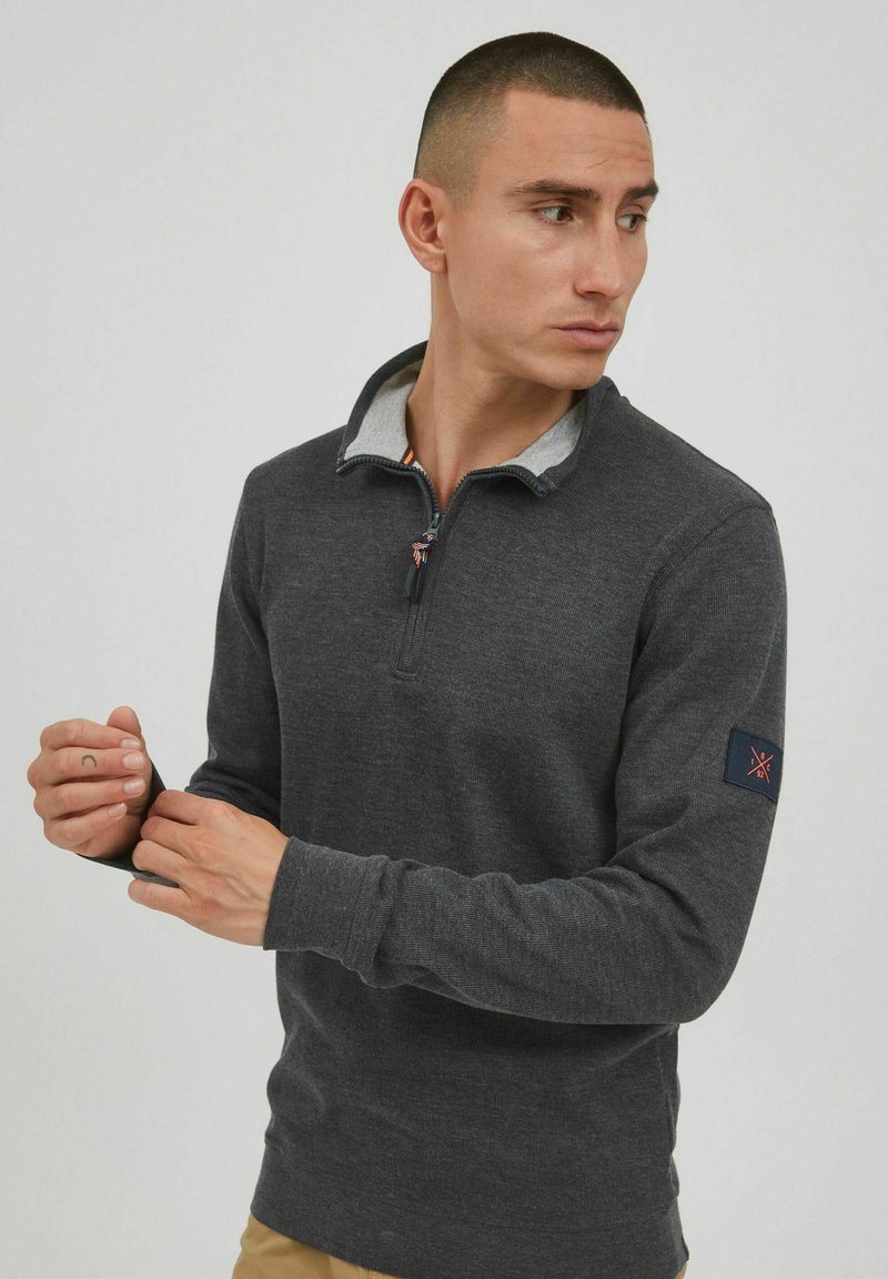 Grå långärmad pullover med dragkedja i kragen, med en grå innervolym och en logopatch på vänster ärm. Slät tygstruktur.