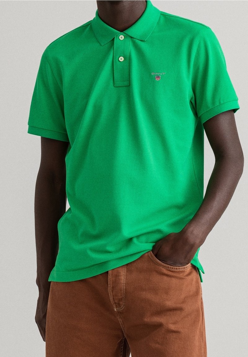 Homme portant un polo vert vif à manches courtes avec col et boutons, associé à un pantalon marron, main dans la poche, fond neutre.