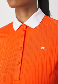 Orange ribbad pikétröja med vit krage, med en knappslå och en liten vit logotyp på vänster bröst.