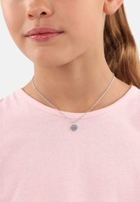 Silver kedja halsband med ett hjärtformat hänge med ett mönster, bärs över en ljusrosa skjorta. Slät textur och minimalistisch design.