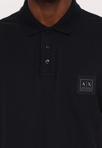 Tumes sinine polo särk, mis on valmistatud tekstureeritud kangast, klassikalise kraega, kolme nööbiga nööpplank ja ruudukujuline logotempel rinnal.