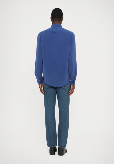 Mann som står med ryggen til, iført en langermet blå skjorte, rette ben blå jeans og svarte sko mot en ensfarget hvit bakgrunn.