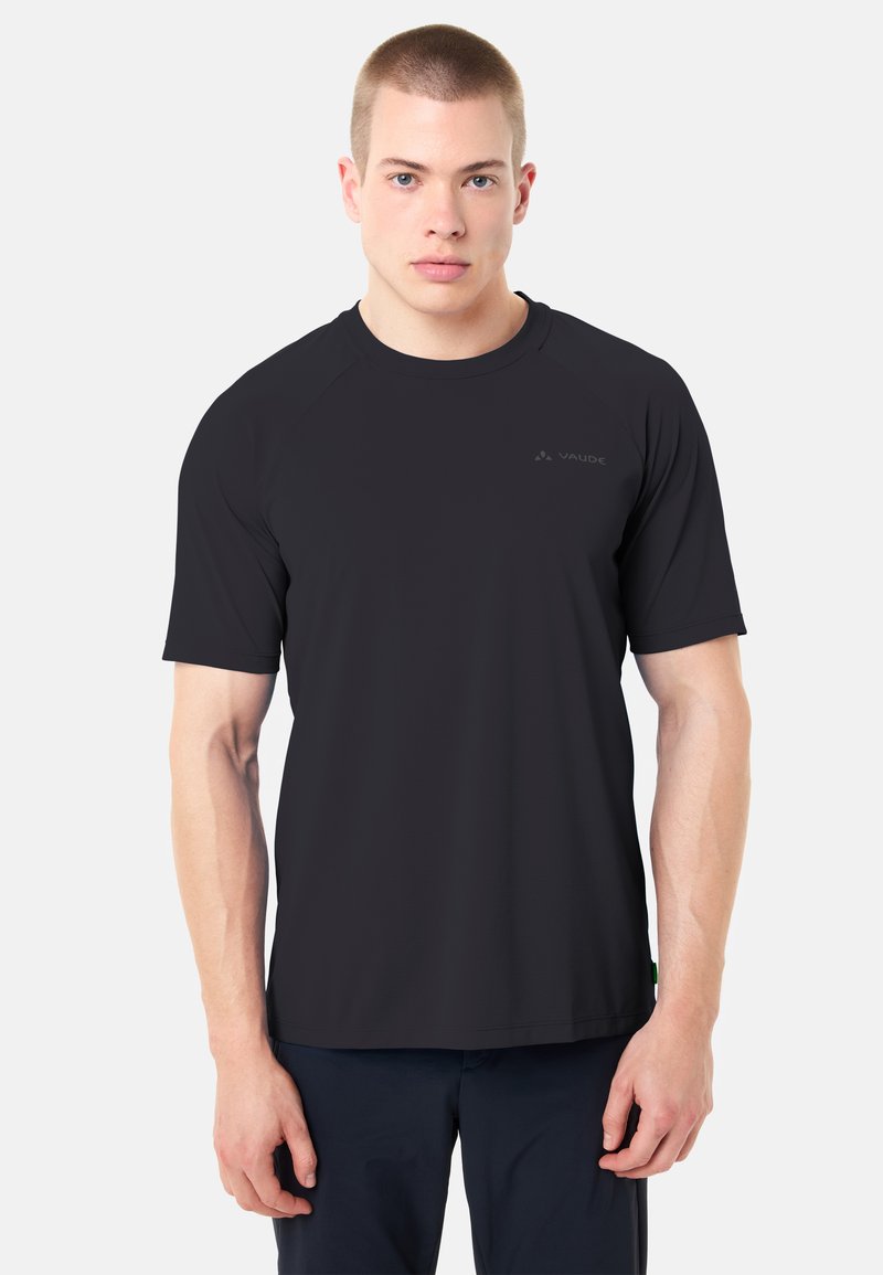 Jeune homme aux cheveux courts portant un t-shirt noir uni à manches courtes et un pantalon foncé, debout devant un fond blanc
