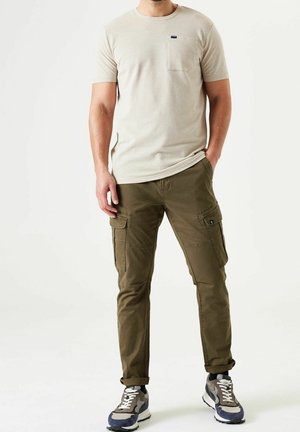 Uomo con t-shirt beige a maniche corte, pantaloni cargo verde oliva e sneakers blu-grigie, in piedi con una mano in tasca.