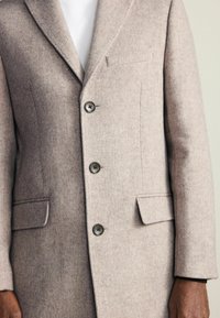 Jasnobeżowy płaszcz z wełny z klapami, trzema guzikami i bocznymi kieszeniami. Gładka struktura z subtelnymi odcieniami kolorów.