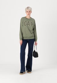 Felpa verde con scritta "GAP", jeans a zampa d'elefante blu scuro, borsa nera con tracolla a catena e scarpe chiare. Tessuto dalla superficie liscia.