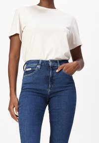 Lyhythihainen, vaalean beige t-paita, jossa on "Calvin Klein" -logo rinnassa, yhdistettynä korkeavyötäröisiin tummansiniisiin farkkuihin.