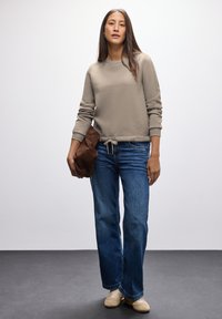 Felpa beige con polsini a costine e orlo con coulisse, abbinata a jeans a gamba larga blu e sneakers di colore chiaro. Il modello tiene una borsa marrone.