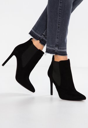 Bottes noires en daim à ankle avec un bout pointu, un talon aiguille haut et des panneaux latéraux élastiques. Portées avec un jean foncé à ourlet effiloché.