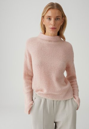 Vrouw met blond haar die een lichtroze gebreide trui en lichtbeige broek draagt, staande tegen een effen grijze achtergrond.