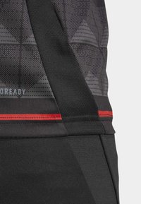 Vêtement de sport noir avec un tissu à motifs aux designs géométriques, une bande d'accent rouge et des textures contrastées le long des coutures.