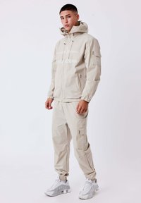 Joven de pie, vistiendo una chaqueta con capucha beige y pantalones cargo a juego con zapatillas blancas, frente a un fondo blanco liso.
