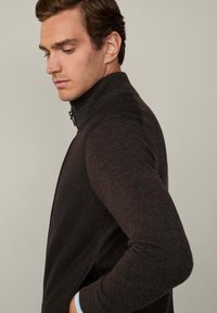 Veste en polaire brun à fermeture éclair avec col montant, présentant un tissu texturé et des manches ajustées. Coutures latérales visibles pour un ajustement sur mesure.