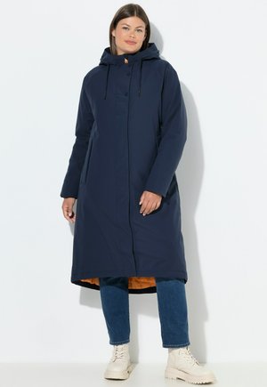 Marineblauer wasserdichter langes Mantel mit abnehmbarem Kapuzenschild, Druckknöpfen, Seitentaschen und einem orangefarbenen Innenfutter. Ergänzt durch beigefarbene Knöchelstiefel.