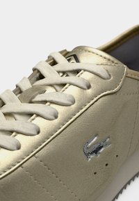 Χρυσά αθλητικά παπούτσια με λείο δερμάτινο επάνω μέρος, λευκά κορδόνια και λογότυπο κροκόδειλου. Διαθέτουν ραμμένες λεπτομέρειες και υφή στην σόλα.