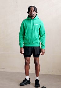 Felpa verde in cotone con tasca frontale, indossata sopra pantaloni corti sportivi neri. Il modello si trova all'interno, indossando scarpe da ginnastica nere e calzini bianchi.