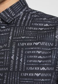 Camiseta negra con un patrón repetido de "Emporio Armani" en blanco, con tamaños y estilos de letra variados. El material parece suave con un cuello estructurado.