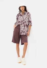 Korte mouwen overhemd in bordeauxrood met een wit tropisch bladpatroon, gecombineerd met knielange shorts in een bijpassende tint en witte sneakers.