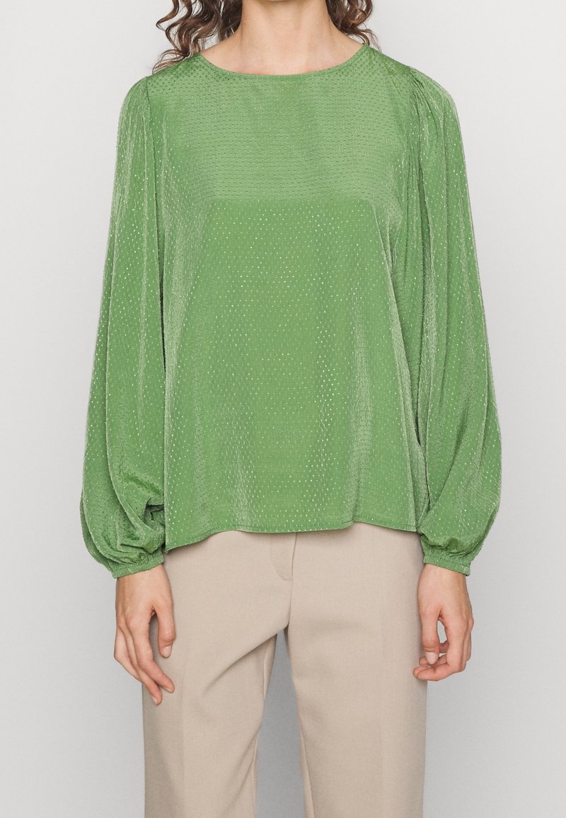 Blouse verte texturée avec des manches longues bouffantes et un col rond. Présente de petits motifs de pois discrets. Associée à un pantalon beige clair.