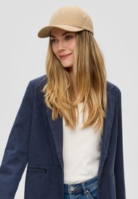 Beige honkbalpet met geborduurd logo, glad materiaal, gebogen rand, gedragen met een marineblauwe blazer over een witte top en blauwe jeans.