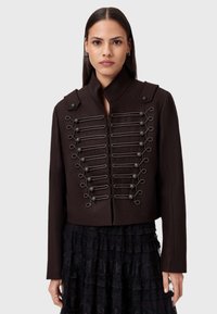 Chaqueta marrón corta con tela texturizada, cuello alto y bucles decorativos con botones en la parte delantera. Combinada con una falda negra con volantes.