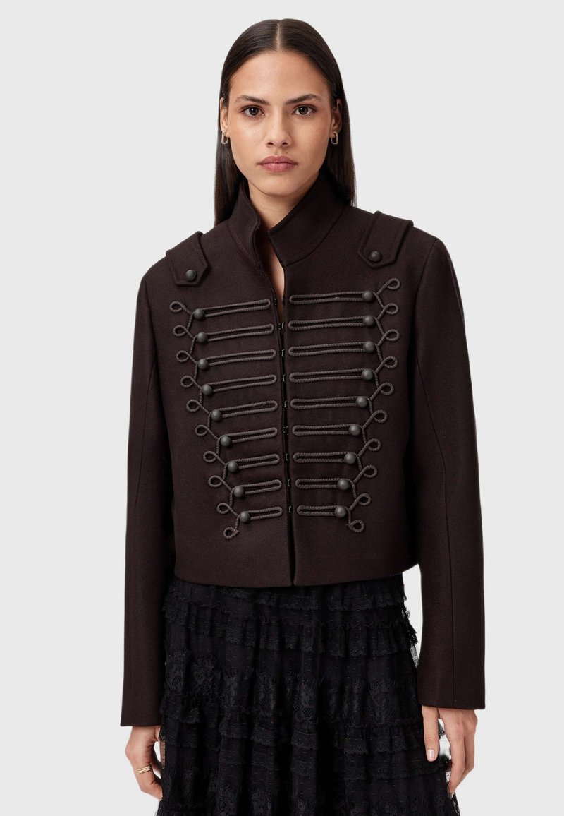 Chaqueta marrón corta con tela texturizada, cuello alto y bucles decorativos con botones en la parte delantera. Combinada con una falda negra con volantes.