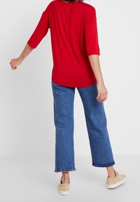 T-shirt rossa oversize con maniche a tre quarti, abbinata a jeans larghi blu chiaro. I jeans presentano un orlo grezzo e sono leggermente strappati.