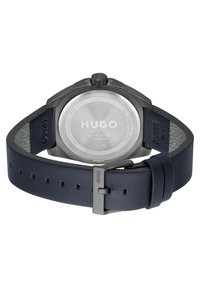Reloj de acero inoxidable gris oscuro con caja redonda, correa de cuero azul marino, esfera circular y fondo texturizado. Presenta el grabado "HUGO" en la caja.