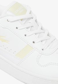 Zapatilla blanca con parte superior de cuero texturizado, con una franja de acento amarilla y un logo cosido. Cordones blancos y suela de goma plana.