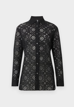 Kvinde iført en sort langærmet blondebluse, sorte vidde bukser, sorte spidse sko, der holder en sammenfoldet rød clutch-taske.