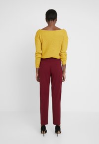 Pull en maille jaune avec un design décolleté porté avec un pantalon ajusté bordeaux et des chaussures à talons noires, vu de derrière.