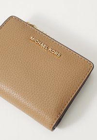 Cartera de cuero color canela con una superficie texturizada, que presenta un logo dorado de "MICHAEL KORS" y un cierre con cremallera. Costuras visibles en los bordes.