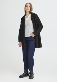 Cappotto lungo nero su una camicia a righe bianche e blu navy, abbinato a jeans blu scuro e stivaletti neri alla caviglia con dettaglio a fibbia. Design minimalista.